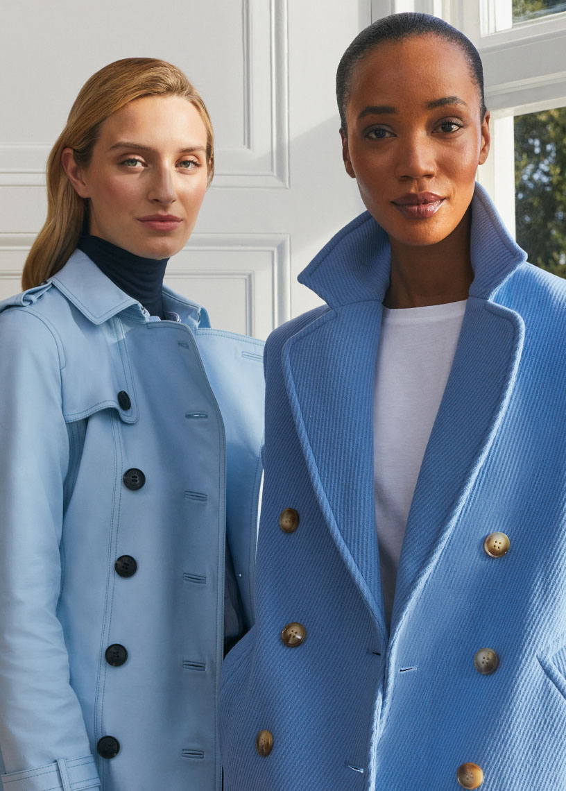 Hobbs models stood by a window wearing a blue trench coat and blue pea coat.